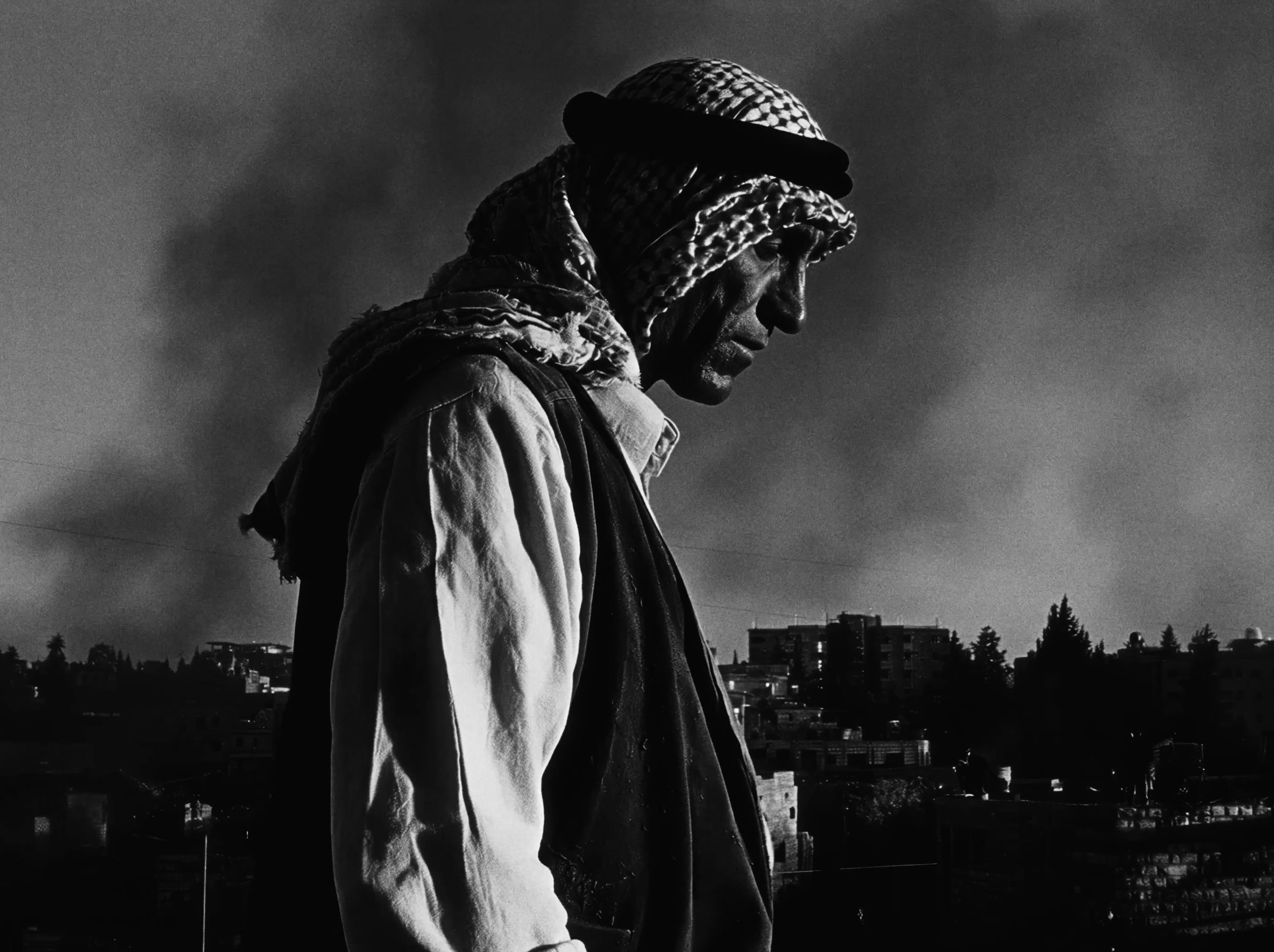 An old man in a keffiyeh before smoke rising over the village.